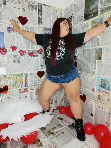 Woman in black graphic tee and denim shorts posing joyfully amidst red balloons and newspaper wall decor.