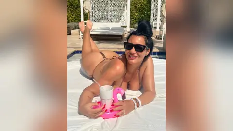 MissyJolie lies on her stomach on a white bed, wearing black sunglasses and a thin black string bikini.