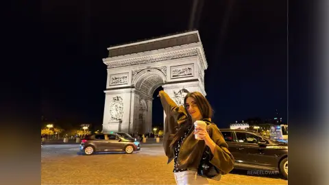 Stylish woman in a brown jacket posing at night in front of the illuminated Arc de Triomphe in Paris.