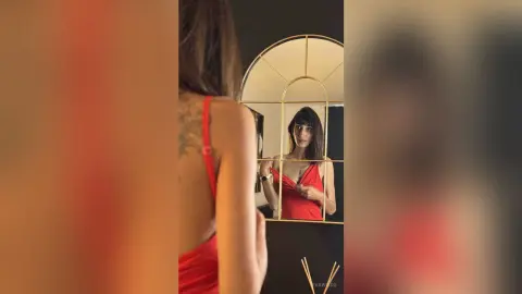 Young woman in a red sleeveless dress taking a mirror selfie in a modern, moody interior setting.