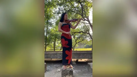 Elegant woman in a red floral maxi dress playing violin in a peaceful green park setting.