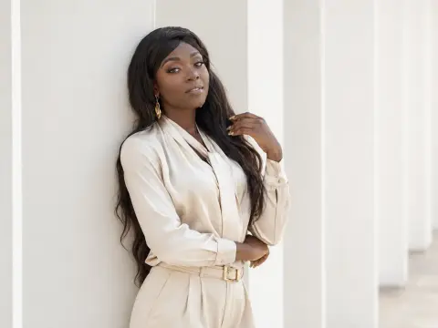 Elegant Black woman in a cream silk blouse leaning against white pillars in a bright, minimalist setting.