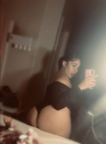 Curvy woman in a black long-sleeve bodysuit taking a moody mirror selfie in a dimly lit room.