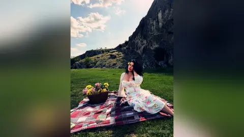 Woman in a white floral dress enjoying a peaceful picnic on a plaid blanket near a rocky mountain cliff.
