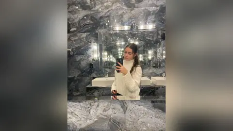Young woman in a white turtleneck taking a mirror selfie in a luxury marble bathroom.
