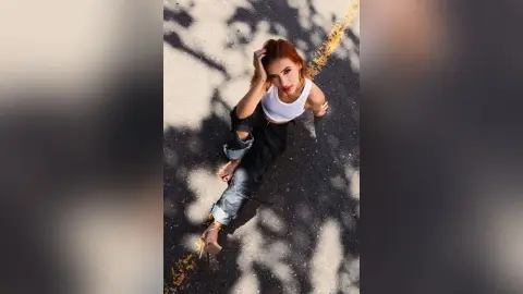 Redheaded woman in a white tank top and ripped jeans sitting on an asphalt road in bright sunlight.