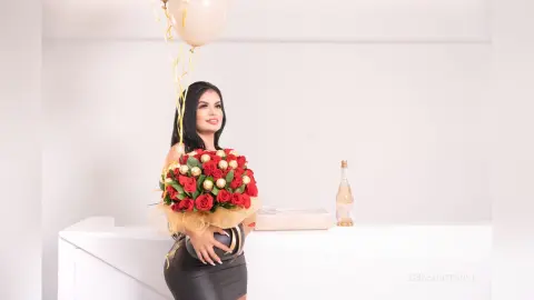 ErikaHoffman smiles while holding a large bouquet of red roses and wearing a tight black dress against a white background.