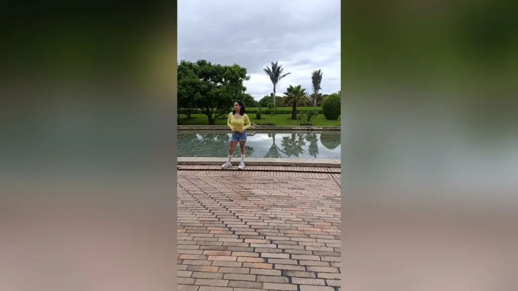 KandyPariston — Young woman in a yellow sweater and denim shorts standing by a serene garden pond under a cloudy sky.