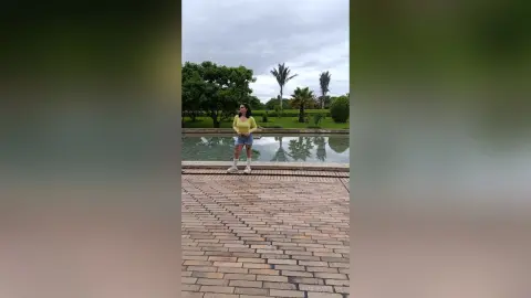 Young woman in a yellow sweater and denim shorts standing by a serene garden pond under a cloudy sky.