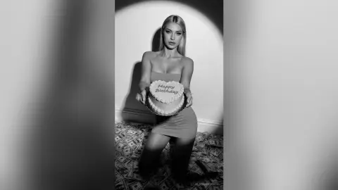 Blonde woman in a sleek strapless dress holding a Happy Birthday cake in a moody black and white studio setting.
