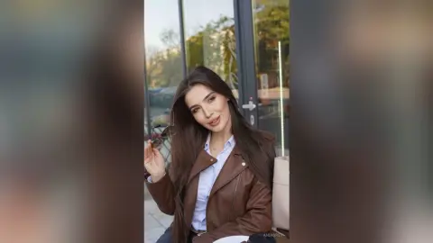 Smiling brunette woman in a brown leather jacket and white shirt sitting outdoors in soft sunlight.