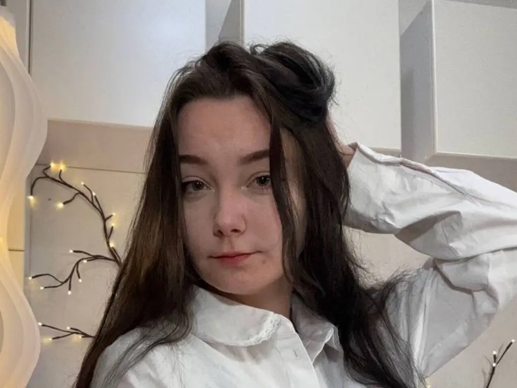Young woman with long brown hair wearing a white button-down shirt in a bright, minimalist room.