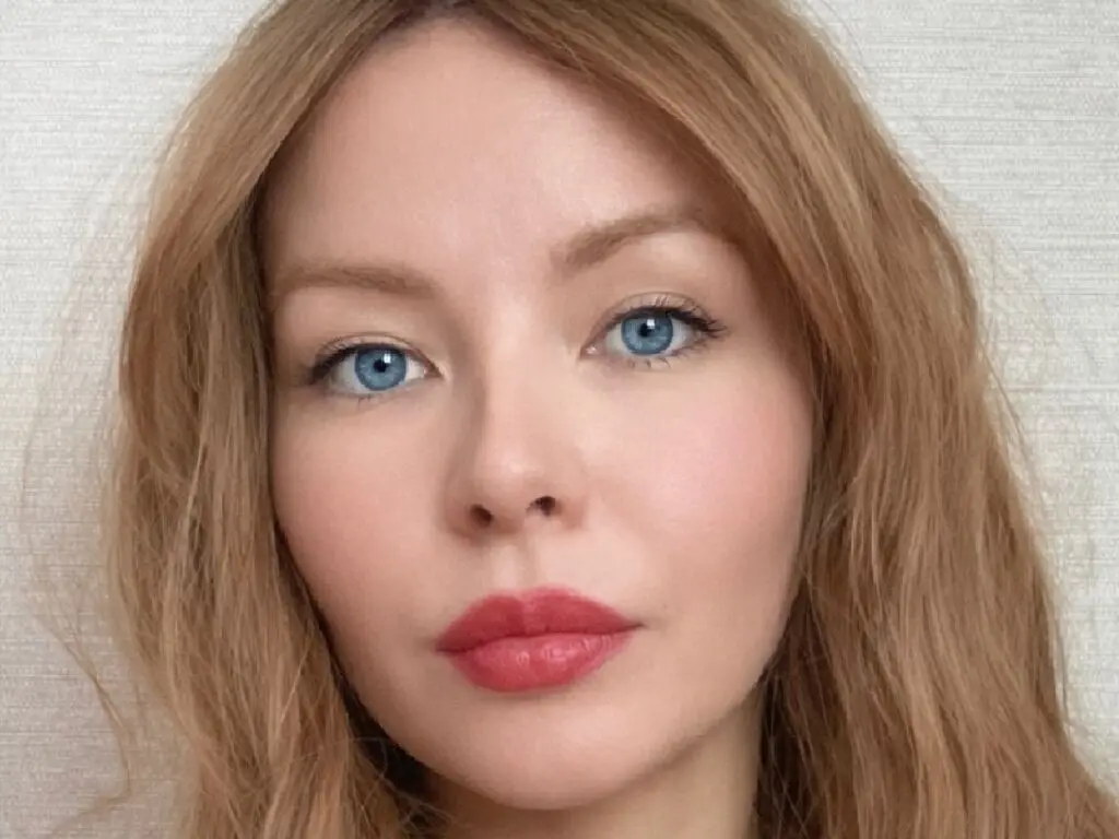 Close-up portrait of a blonde woman with blue eyes and red lipstick looking serene against a neutral background.