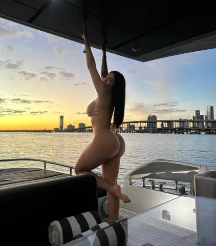 Slim woman in a bikini relaxing on a luxury yacht at sunset overlooking a city skyline.