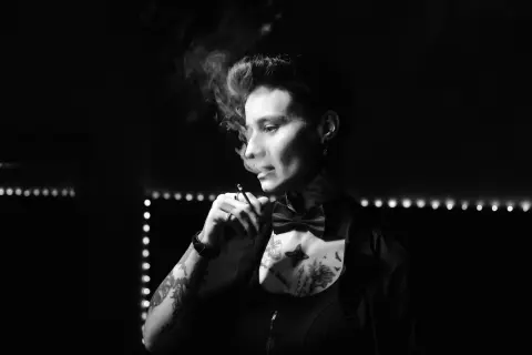 Moody black and white portrait of a tattooed person in a bow tie smoking in a dimly lit lounge.