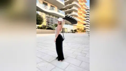 KaylaBryan stands on a sunny balcony wearing a white cap, a tan crop top, and black pants.