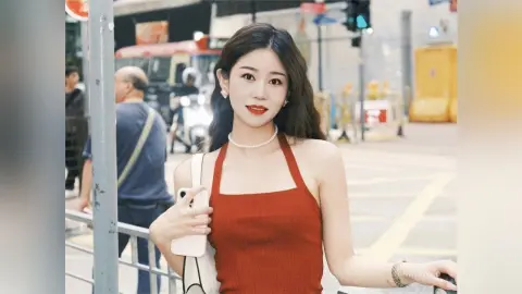Young woman in a red halter top and pearl necklace smiling on a sunny city street.