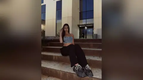 Young woman in denim tube top and black trousers sitting on outdoor stairs at night.
