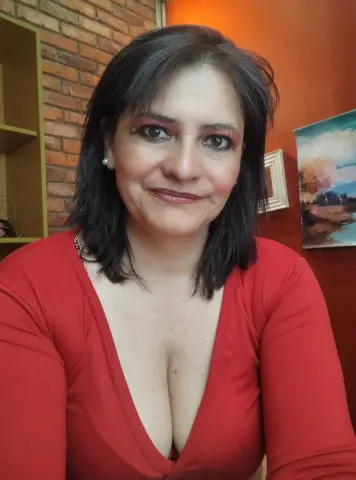 Smiling woman in a red V-neck top sitting in a cozy room with brick walls and warm lighting.