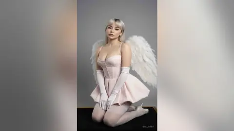 Blonde woman in a pink angel costume with white wings and gloves posing elegantly against a grey background.