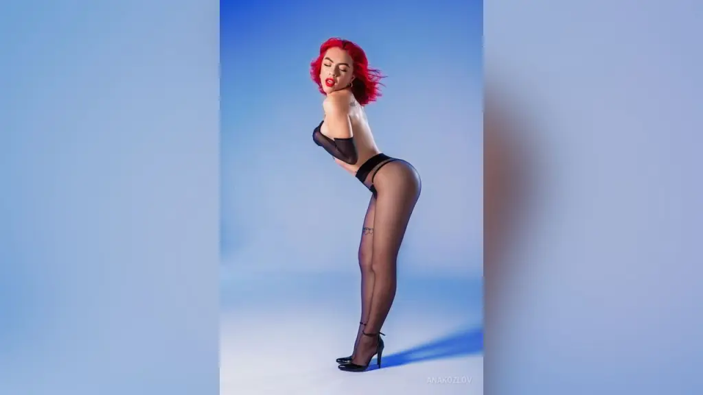 AnaKozlov β Red-haired woman in black lingerie and sheer tights posing confidently against a blue studio background.
