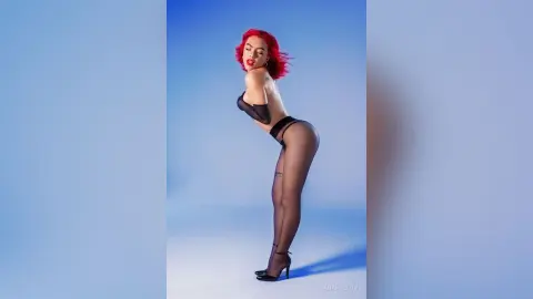 Red-haired woman in black lingerie and sheer tights posing confidently against a blue studio background.