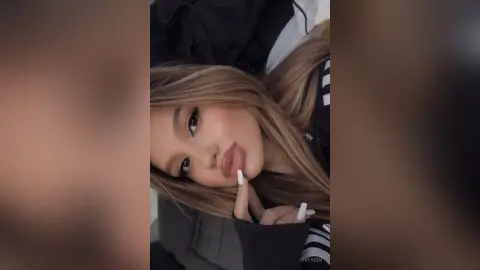AnyaSin poses with a hand near her lips, showing her long blonde hair and heavy eye makeup.