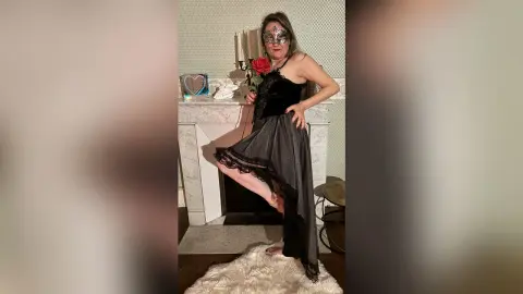 Woman in a black lace masquerade dress holding a red rose by a marble fireplace in a moody setting.