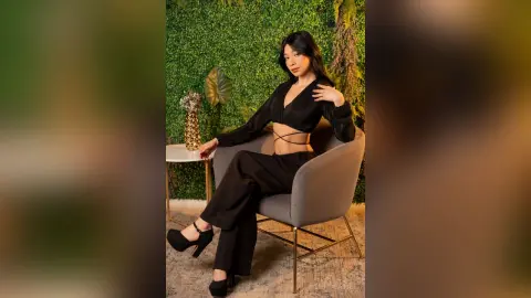 Elegant woman in a black crop top and trousers sitting in a grey chair against a lush green leaf wall.