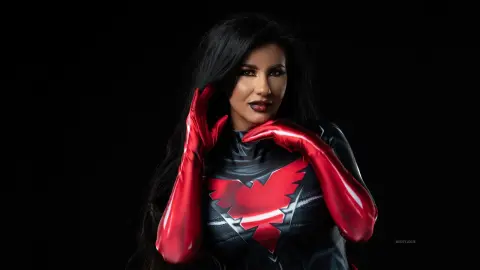 MissyJolie poses in a black and red superhero bodysuit with long red gloves against a black background.