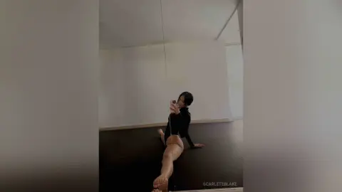 Woman in black bodysuit stretching on a dark floor in a minimalist white room, moody aesthetic.