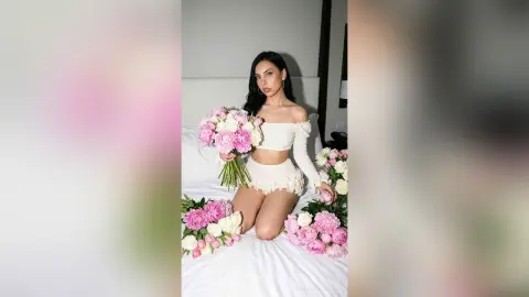 Young woman in a white off-the-shoulder crop top sitting on a bed surrounded by pink peony bouquets.