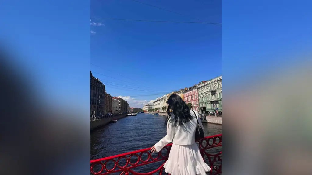 SofiMils β SofiMils stands by a canal in a white long-sleeved dress with her long dark hair flowing against a clear blue sky.