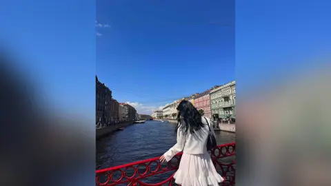 Woman in a white dress standing by a canal in a European city under a clear blue sky.