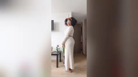 Smiling woman in a white ribbed knit maxi dress and sunglasses standing in a bright modern living room.