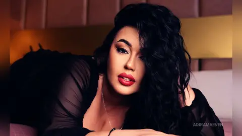 AdiraMaeven poses with her long, wavy black hair, wearing a sheer black top and red lipstick against a warm, golden headboard.