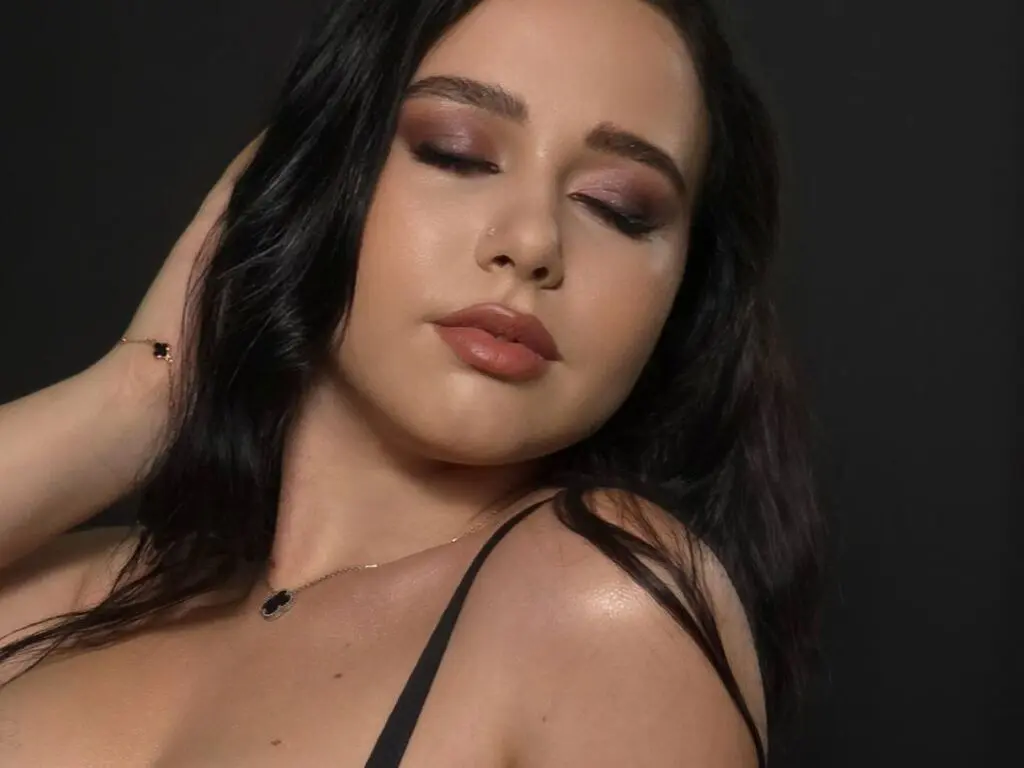 Woman with dark wavy hair and smoky eye makeup wearing a black strap against a dark studio background.
