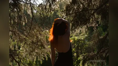 Back view of a woman in a black tank top standing under sunlit trees in a dreamy forest setting.