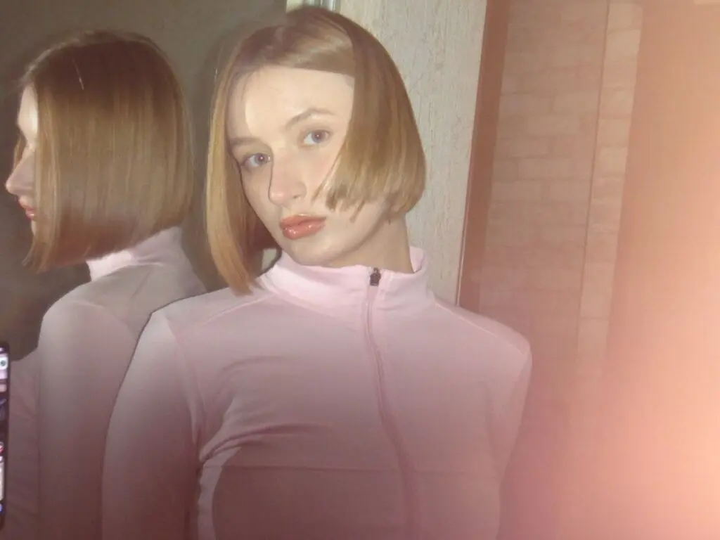 Young woman with a blonde bob wearing a pink zip-up sweatshirt in a soft, warm indoor setting.