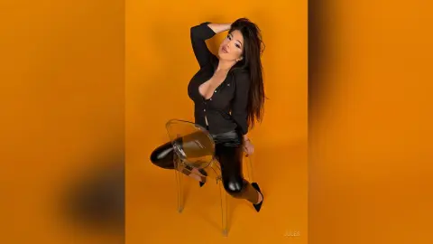 Glamorous woman in a black button-down shirt and leather pants sitting on a clear chair against an orange background.