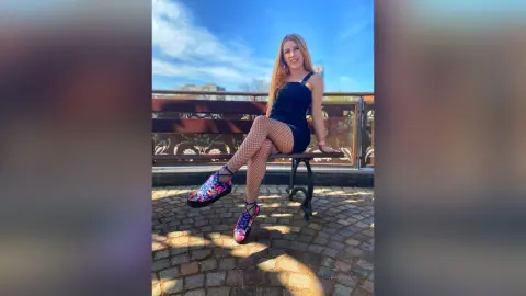 Smiling woman in a black mini dress and patterned tights sitting on a rooftop bench under a blue sky.