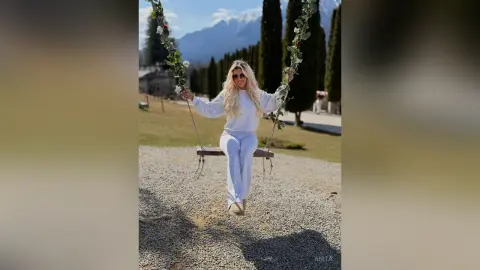 Anita sits on a wooden swing wearing a white long-sleeved top and white pants against a mountain backdrop.