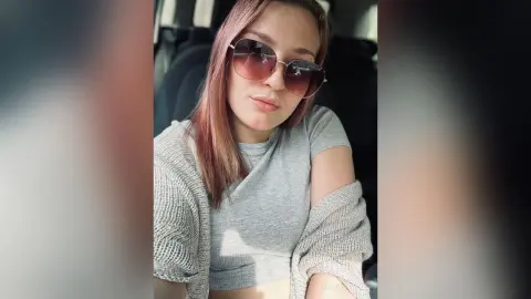 CristaStwell wearing a grey t-shirt and a light grey knit cardigan while sitting in a car, looking at the camera through large sunglasses.
