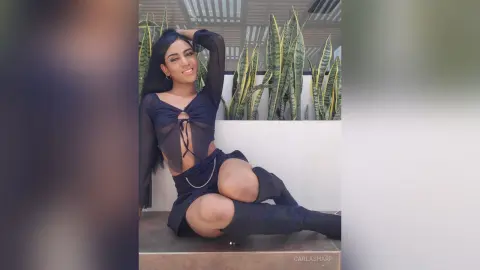 Smiling woman in a black sheer long-sleeve top and mini skirt sitting outdoors by tall snake plants.