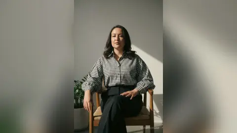 Confident woman in a black and white checkered shirt sitting in a minimalist studio setting.