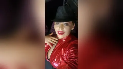 DonatellaRossy — DonatellaRossy wears a black fedora and a shiny red jacket while smiling at the camera.