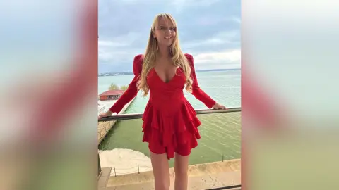 Smiling blonde woman in a stylish red cocktail dress standing on a balcony overlooking the ocean.