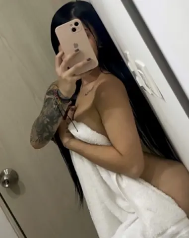 Young woman with long dark hair wrapped in a white towel taking a mirror selfie in a bright bathroom.