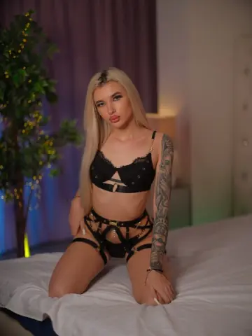 Juicy_Jessye poses on a white bed wearing black lace lingerie and a garter belt, showing off her blonde hair and arm tattoos.