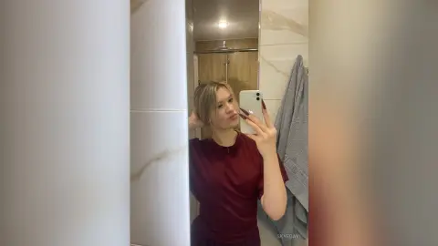 Young woman in a maroon t-shirt taking a casual mirror selfie in a modern bathroom.